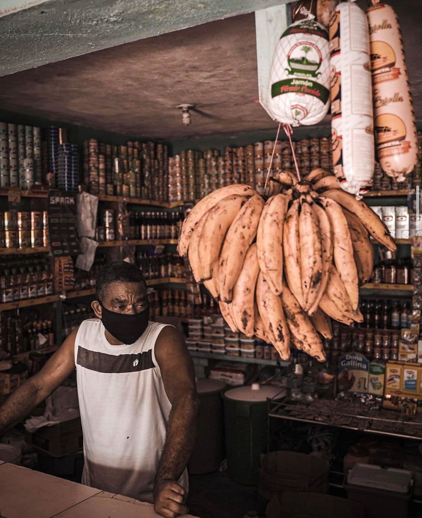 A shopkeeper behind the counter at the corner store, shelves stocked with canned goods and plantains overhead