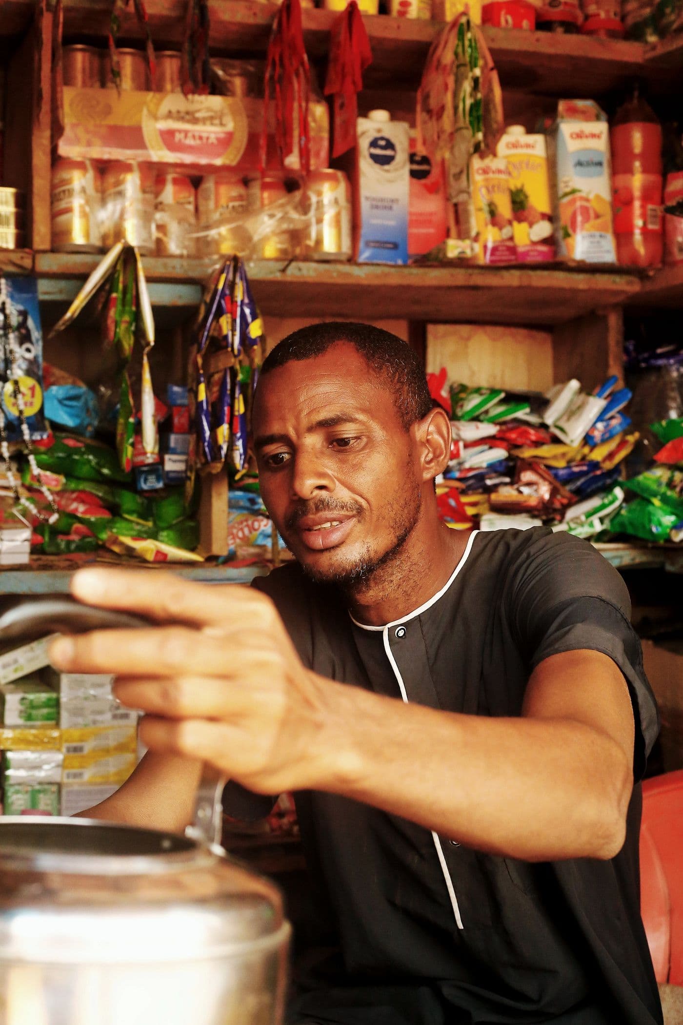 A shopkeeper reaching across the counter, snacks and drinks stocked on the shelves behind
